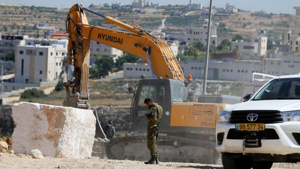 A member of Israeli security forces uses a mobile phone as a machinery removes stone blocks placed by Palestinians in Hebron in the Israeli-occupied West Bank July 2, 2020.  - Sputnik International