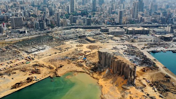 An aerial view taken on August 7, 2020, shows a partial view of the port of Beirut, the damaged grain silo and the crater caused by the colossal explosion three days earlier of a huge pile of ammonium nitrate that had languished for years in a port warehouse, leaving scores of people dead or injured and causing devastation in the Lebanese capital. The city of Beirut can be seen in the background. - Sputnik International