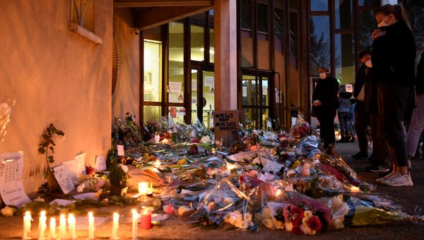 People hold a vigil and lay flowers and candles next to a placard reading 'I am a teacher, I am Samuel' at the entrance of a middle school in Conflans-Sainte-Honorine, 30kms northwest of Paris, on 17 October 2020. History teacher Samuel Paty was beheaded by an attacker after showing his pupils cartoons of the prophet Mohammed. The man suspected of the murder on 16 October 2020 was an 18-year-old who had been born in Moscow and originated from Russia's southern region of Chechnya, a judicial source said on 17 October. Five more people have been detained over the murder including the parents of a child at the school where the teacher was working, bringing to nine the total number currently under arrest, said the source, who asked not to be named. The attack happened at around 5 pm (1500 GMT). - Sputnik International