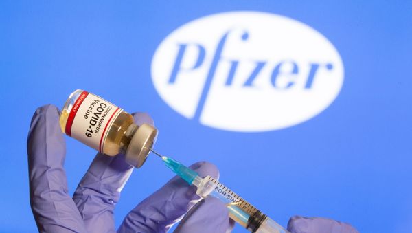 A woman holds a small bottle labelled with a Coronavirus COVID-19 Vaccine sticker and a medical syringe in front of displayed Pfizer logo in this illustration taken, October 30, 2020. - Sputnik International