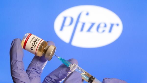 A woman holds a small bottle labeled with a Coronavirus COVID-19 Vaccine sticker and a medical syringe in front of displayed Pfizer logo in this illustration taken, October 30, 2020.  - Sputnik International