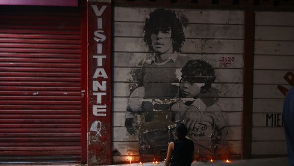 A boy kneels in front of a mural of Diego Maradona decorated with canddles outside the stadium of Argentinos Juniors soccer club, where he started as a professional footballer, in Buenos Aires, Argentina, Wednesday, Nov. 25, 2020. The Argentine soccer great who was among the best players ever and who led his country to the 1986 World Cup title died from a heart attack on Wednesday at his home in Buenos Aires. He was 60.  - Sputnik International