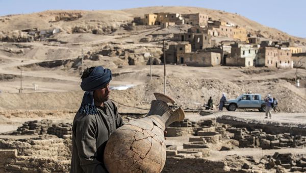 A picture taken on April 10, 2021, shows a worker carrying a pot at the archaeological site of a 3000 year old city, dubbed The Rise of Aten, dating to the reign of Amenhotep III, uncovered by the Egyptian mission near Luxor. - Archaeologists have uncovered the remains of an ancient city in the desert outside Luxor that they say is the largest ever found in Egypt and dates back to a golden age of the pharaohs 3,000 years ago. Famed Egyptologist Zahi Hawass announced the discovery of the lost golden city, saying the site was uncovered near Luxor, home of the legendary Valley of the Kings.  - Sputnik International