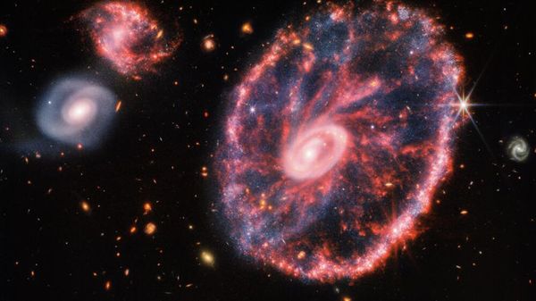 A large pink, speckled galaxy resembling a wheel with with a small, inner oval, with dusty blue in between on the right, with two smaller spiral galaxies - Sputnik International