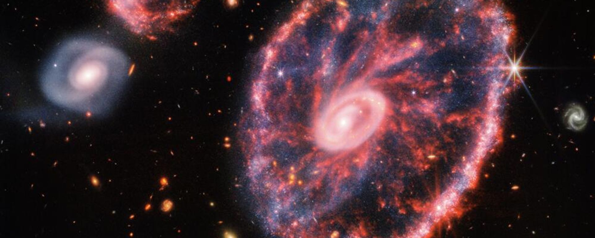 A large pink, speckled galaxy resembling a wheel with with a small, inner oval, with dusty blue in between on the right, with two smaller spiral galaxies - Sputnik International, 1920, 13.09.2023