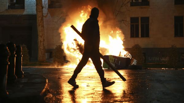 A man with a stick in hand walks past a burning police car not far from the Georgian parliament building in Tbilisi, Georgia, Thursday, March 9, 2023. - Sputnik International