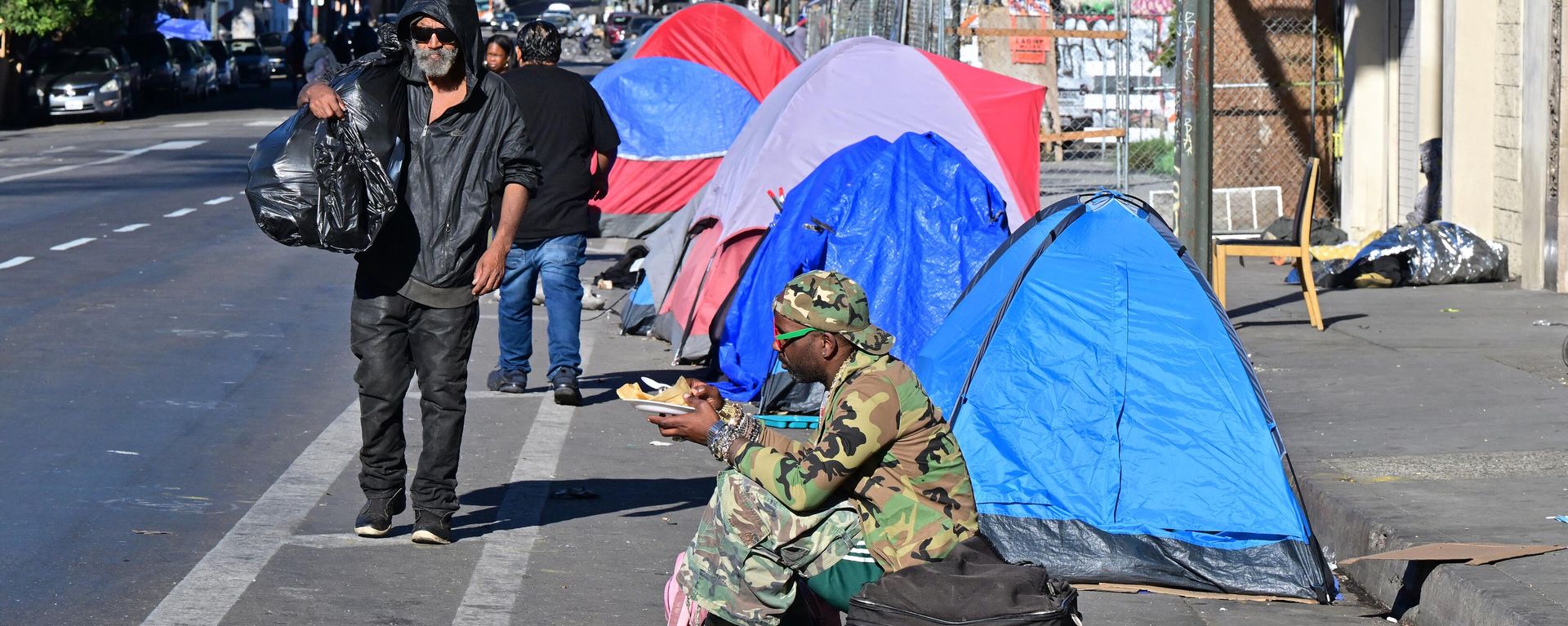 A homeless man sits on a suitcase on the side of a street lined with tents housing the homeless in Downtown Los Angeles on November 22, 2023. - Sputnik International, 1920, 27.12.2023