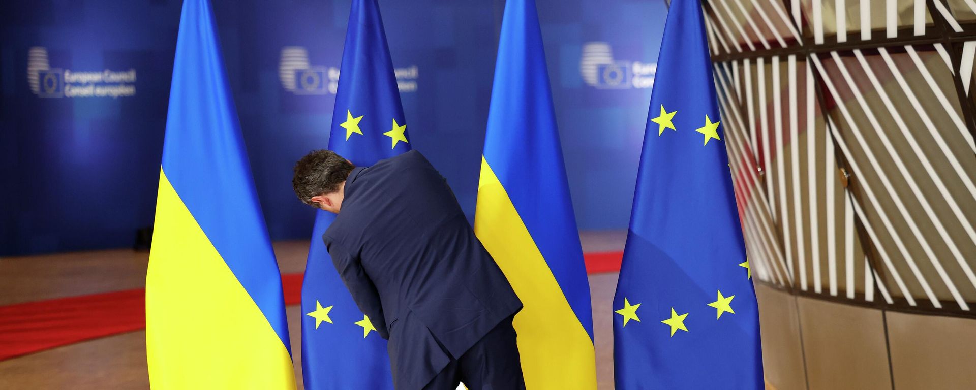 A member of protocol adjusts the EU and Ukrainian flags prior to arrivals for an EU summit at the European Council building in Brussels, Dec. 19, 2024. - Sputnik International, 1920, 16.04.2026