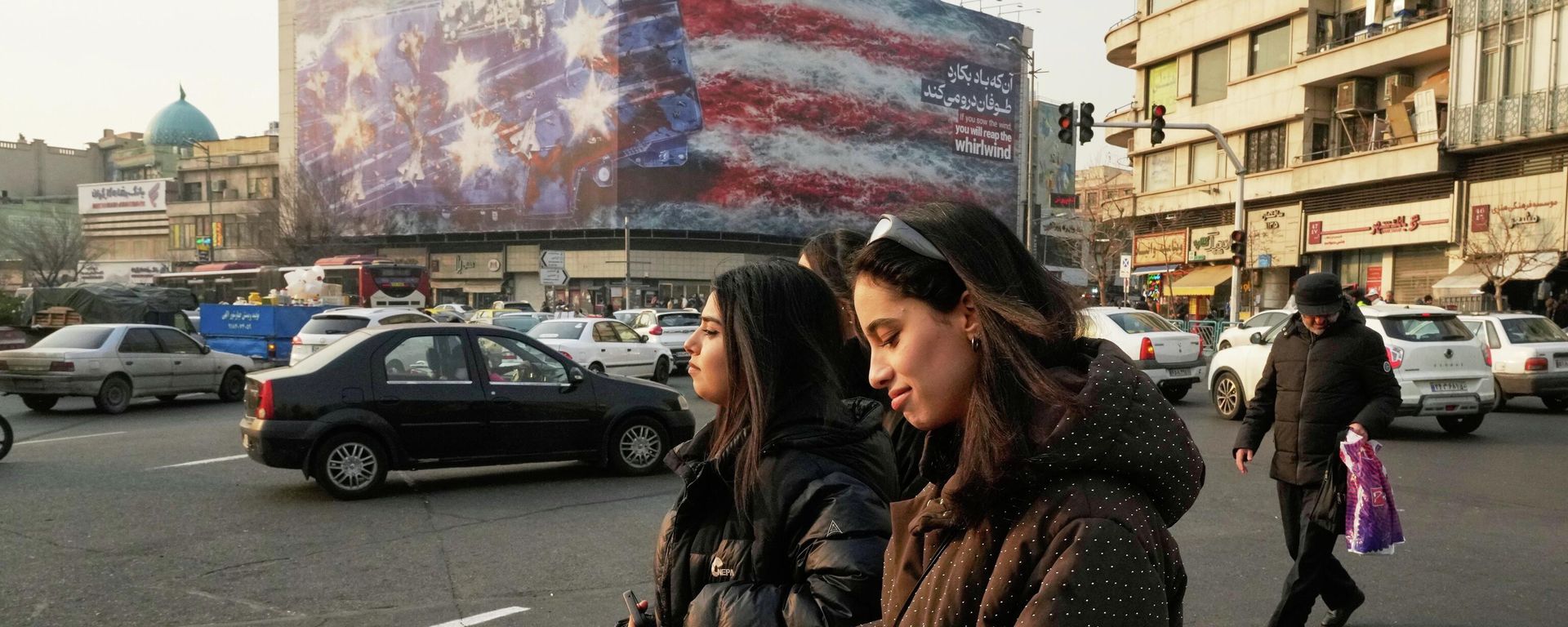 People walk past a billboard depicting a damaged U.S. aircraft carrier with disabled fighter jets on its deck and a sign reading in Farsi and English, If you sow the wind, you'll reap the whirlwind, at Enqelab-e-Eslami (Islamic Revolution) Square in Tehran, Iran, Sunday, Jan. 25, 2026. - Sputnik International, 1920, 09.04.2026
