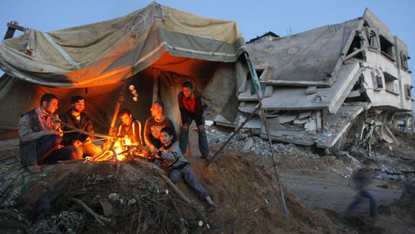 Palestinians sit around a fire under the cover of a tent on the ruins of their home Palestinians sit around a fire under the cover of a tent on the ruins of their home - Sputnik International