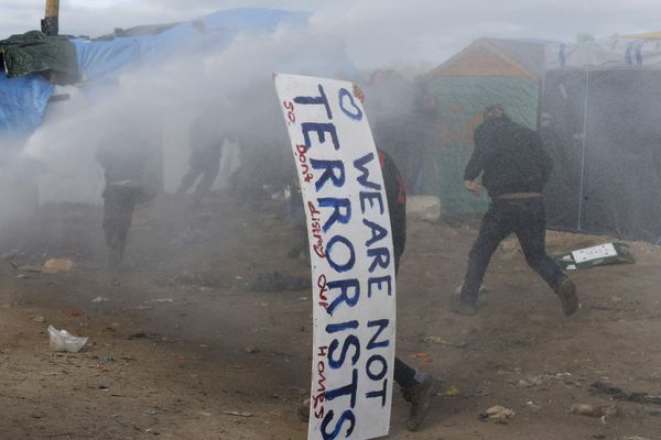A man takes cover with a sign that reads, We Are Not Terrorists - So Don't Destroy Our Homes as water canon and tear gas push back migrants. - Sputnik International
