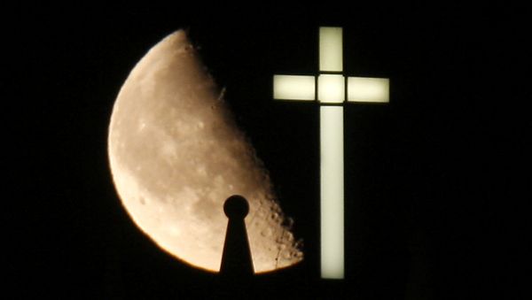 A third quarter Buck Moon rises behind a spire on the top of the bell tower next to a lighted cross - Sputnik International