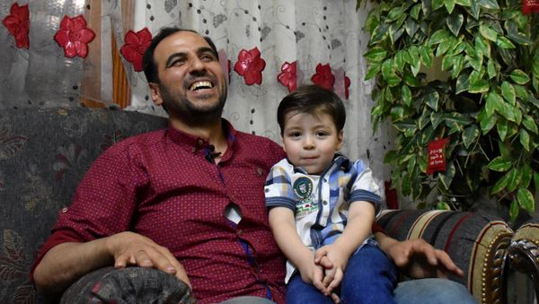 A picture taken on June 6, 2017, shows Omran, a Syrian boy pictured last year in an ambulance after being rescued from the rubble of a building, sitting on the lap of his father at their new home in Aleppo - Sputnik International