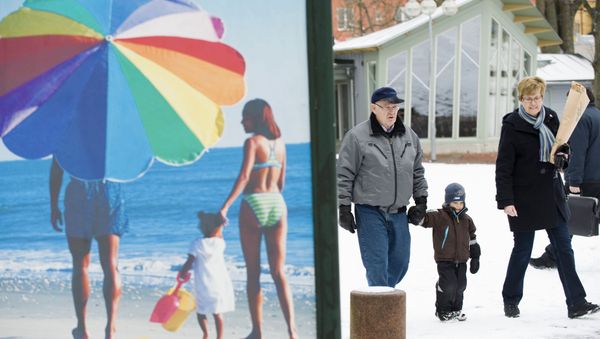 People walk along a snow covered street past an advertisement featuring a pictures of a beach scene in Mariehamn. (File) People walk along a snow covered street past an advertisement featuring a pictures of a beach scene in Mariehamn. (File) - Sputnik International