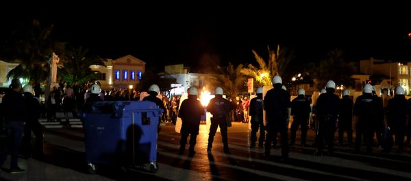 A garbage bin burns as riot police officers stand guard separating protesting groups of locals and refugees demonstrating against conditions in Moria camp and delays in asylum applications, in the city of Mytilene on the island of Lesbos, Greece, April 22, 2018 A garbage bin burns as riot police officers stand guard separating protesting groups of locals and refugees demonstrating against conditions in Moria camp and delays in asylum applications, in the city of Mytilene on the island of Lesbos, Greece, April 22, 2018 - Sputnik International