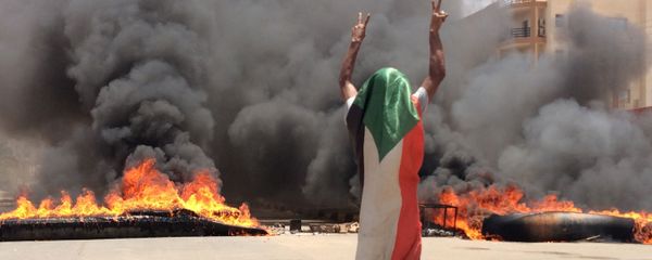 A protester wearing a Sudanese flag flashes the victory sign in front of burning tires and debris on road 60, near Khartoum's army headquarters. - Sputnik International