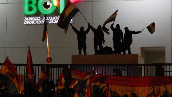 Police officers standing on the rooftop of a security booth at a police station wave Bolivian flags in front of protesters in La Paz - Sputnik International