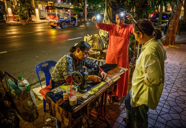 A woman uses the lights of two mobile phones to help a street tailor finish her work on a street in Bangkok on December 3, 2019. A woman uses the lights of two mobile phones to help a street tailor finish her work on a street in Bangkok on December 3, 2019. - Sputnik International